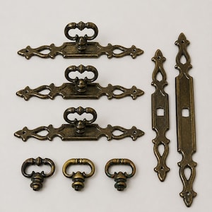 May include: A collection of antique brass-toned metal hardware including three horizontal drawer pulls with ornate backplates, three matching drop-style knobs, and two vertical decorative plates with central square openings for mounting.