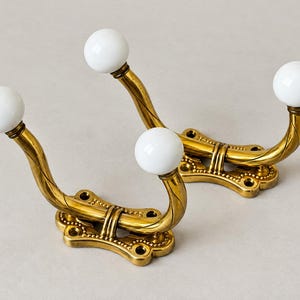 May include: Two wall-mounted coat hooks made of polished brass with a twisted metal arm design. Each hook features a smooth, round white ceramic ball at the tip and a rectangular base plate with four pre-drilled screw holes for installation.