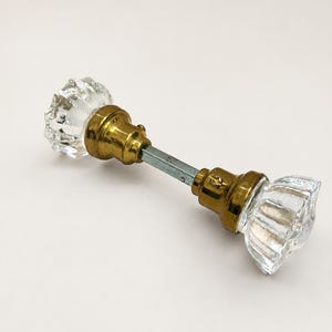 May include: A door knob set featuring two clear, fluted glass knobs attached to a central brass-toned metal base and a square metal spindle connecting the two sides. The design includes a metallic collar at the base of each glass knob.