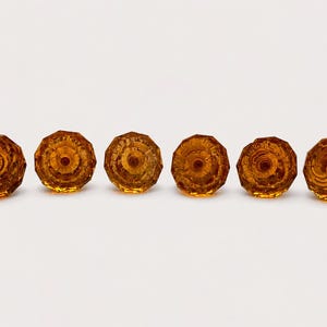 May include: A set of six amber-colored glass knobs, each featuring a faceted, multi-sided circular design with a central hole. The knobs are arranged in a horizontal row, showcasing a translucent, warm orange hue and a reflective surface.