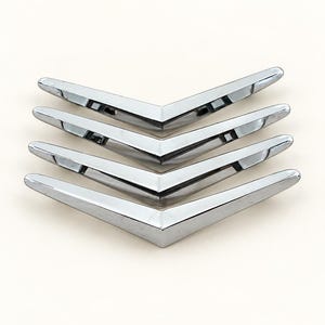 May include: A set of four identical chevron-shaped metal emblems with a polished chrome finish. Each piece features a sharp V-angle design with smooth, reflective surfaces and a tapered profile, arranged in a stacked vertical formation.