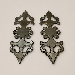 May include: Two matching elongated metal escutcheon plates with a dark bronze finish. Each plate features a symmetrical, ornate scrollwork design with a pointed top and bottom, and includes two small circular holes for mounting hardware.