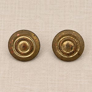 May include: Two circular brass buttons with a weathered, aged patina. Each button features a raised, concentric ring design with a prominent, rounded center dome. The metal surface shows visible signs of oxidation and wear, creating a textured, metallic appearance across the faces of both round fasteners.