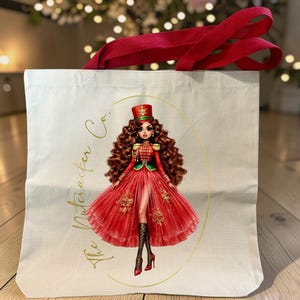 May include: A cream-colored canvas tote bag with red fabric handles. The front features a graphic of a doll with long brown curly hair wearing a red military-style jacket and a red tulle skirt. The text The Nutcracker Co. is printed in gold.
