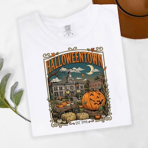 May include: A white t-shirt featuring a graphic print of a town square with a large jack-o'-lantern, a wagon filled with pumpkins, and hay bales. The text reads HALLOWEENTOWN and EST 1998. The shirt has a crew neck and a Comfort Colors brand label.