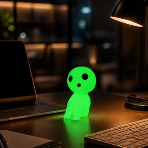 May include: A small, glowing green lamp shaped like a stylized humanoid figure with a large round head, two dark oval eyes, and a small dark mouth. The lamp emits a bright, soft light, illuminating the surface it sits on in a dark room.