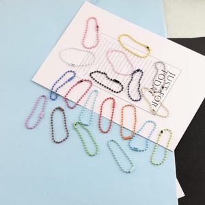 Può includere: Una collezione di piccole catenelle a palline di forma ovale in vari colori, tra cui rosa, giallo, blu, nero, bianco, verde, arancione e viola. Ogni catenella è dotata di una piccola chiusura in metallo. Le catenelle sono disposte su un cartoncino bianco.