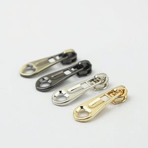 May include: Four metal zip pulls arranged in a diagonal line. Each pull features a teardrop-shaped body with a heart-shaped cutout at the bottom. The set includes one antique brass, one gunmetal, one silver-toned, and one gold-toned finish.