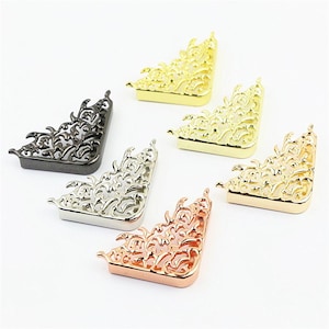 May include: A collection of six decorative metal corner protectors featuring an intricate filigree cutout pattern. The set includes pieces in various metallic finishes, specifically gold, silver, gunmetal black, and rose gold, arranged in a cluster.