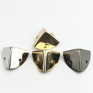 Può includere: Quattro paraspigoli in metallo per scatole o libri, caratterizzati da una forma triangolare curva e liscia. Il set include un pezzo color argento, due color oro e uno color canna di fucile, ciascuno con due piccoli fori per il fissaggio.