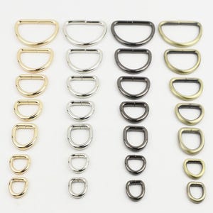 Op de afbeelding: Een verzameling metalen D-ringen gerangschikt in vier verticale kolommen op kleur, waaronder goud, zilver, gunmetal en antiek messing. Elke kolom bevat zeven D-ringen van verschillende groottes, aflopend van de grootste naar de kleinste.
