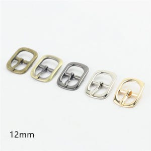 May include: Five metal belt buckles with a 12mm inner width, arranged in a row. The buckles feature different finishes including antique brass, light gold, gunmetal, silver, and polished gold, each with a central prong and rounded frame.