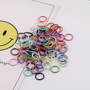 Op de afbeelding: Een verzameling kleine, ronde metalen ringen in diverse levendige kleuren, waaronder rood, blauw, groen, geel, roze, paars, wit en zwart. Deze dunne, open ringen liggen verspreid in een hoop en tonen een gevarieerd kleurenpalet.