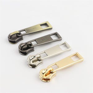 May include: Four metal zipper pulls arranged in a diagonal row. The pulls feature a long, flat rectangular tab with a square cutout at the end and a rounded loop for attachment. The finishes include antique brass, gunmetal, silver, and bright gold.