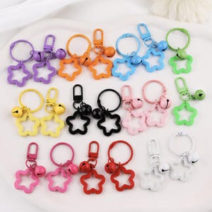 May include: A collection of metal star-shaped keychains in various colors including purple, orange, blue, green, yellow, black, pink, red, and white. Each keychain features a circular ring, a lobster clasp, and a small round jingle bell attached.