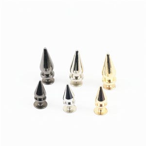 May include: A collection of six metal cone-shaped studs arranged in two rows. The studs feature a pointed top, a rounded base, and a screw-back mechanism. They are finished in three distinct metallic colours: gunmetal grey, polished silver, and gold.