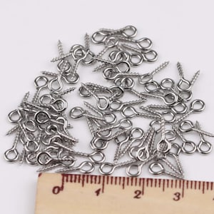 Puede incluir: Una colección de armellas roscadas de metal plateado con vástagos roscados y bucles circulares. Una regla de madera está colocada en la parte inferior, mostrando que las armellas miden aproximadamente 1,3 a 2,5 cm de largo en la escala.