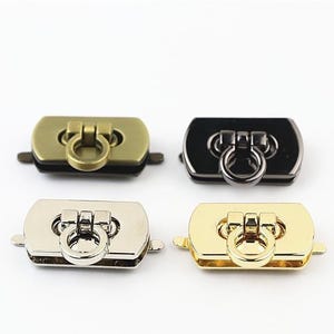 May include: Four rectangular metal bag locks arranged in a grid. Each lock features a central circular ring pull mechanism. The finishes include antique brass, glossy black, polished silver, and bright gold, each with side tabs for attachment.