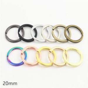 Puede incluir: Una colección de once anillas metálicas circulares para llaves dispuestas en dos filas, cada una de 20 mm de diámetro. Las anillas presentan varios acabados, como negro mate, plata, bronce, arcoíris iridiscente, oro rosa y tonos dorados.