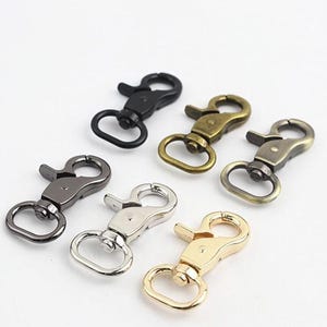 Könnte beinhalten: Eine Sammlung von sechs drehbaren Karabinerverschlüssen aus Metall in verschiedenen Ausführungen, darunter Mattschwarz, Antikmessing, Rotguss, poliertes Silber und glänzendes Gold. Jeder Verschluss hat einen Federriegel und eine drehbare Basis.