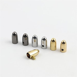 May include: A collection of six small, bullet-shaped metal end caps in various finishes including gunmetal, silver, and gold. Five caps stand upright, while one gold-toned cap lies on its side, revealing a small threaded screw attachment.