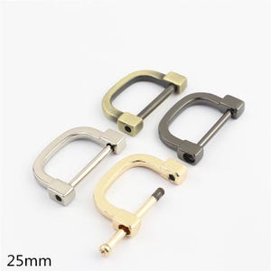 Puede incluir: Cuatro hebillas metálicas en forma de D con un ancho de 25 mm. Tres hebillas están completamente ensambladas con una barra recta, mientras que una hebilla dorada se muestra con su barra roscada desmontada. Acabados en latón antiguo, bronce, plata y oro.