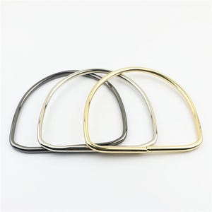 Op de afbeelding: Drie D-vormige metalen tashandvatten in verschillende metallic afwerkingen. Eén handvat is donker gunmetal grijs, één is zilverkleurig en één is goudkleurig. Elk handvat heeft een glad, afgerond frame met een platte onderkant.