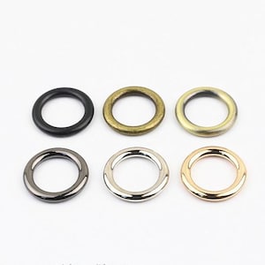 May include: Six circular metal rings arranged in two rows of three. The top row features a matte black ring, an antique bronze ring, and a brushed gold ring. The bottom row includes a gunmetal ring, a polished silver ring, and a rose gold ring.