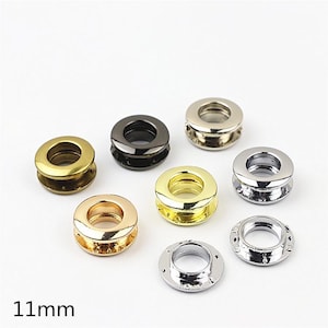 May include: A collection of seven metal grommets in various finishes including gold, silver, gunmetal, and brass. Each circular eyelet features a smooth, rounded rim and a central opening. The text 11mm is printed in black in the bottom left corner.