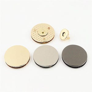 Könnte beinhalten: Eine Sammlung runder magnetischer Taschenverschlüsse in verschiedenen metallischen Ausführungen wie Gold, Silber und Anthrazit. Das Set enthält einen strukturierten goldenen Verschluss mit sichtbaren Schrauben und ein kleineres passendes Magnetteil sowie drei glatte, einfarbige runde Scheiben in Gold-, Silber- und Dunkelgrautönen.