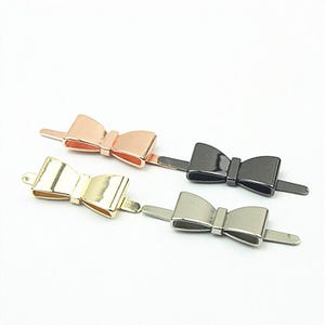 Peut inclure: Quatre attaches métalliques en forme de nœud avec des finitions or rose, or, noir et argent. Chaque attache présente un design de nœud central avec deux tiges plates et allongées de chaque côté pour la fixation, avec une surface métallique lisse.