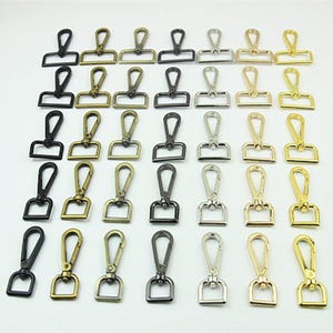 Könnte beinhalten: Eine Sammlung von drehbaren Karabinerverschlüssen aus Metall, die in einem Raster angeordnet sind. Das Set enthält verschiedene Ausführungen wie Mattschwarz, Antikmessing, Gunmetal, Silber und poliertes Gold, jeweils mit einer rechteckigen Basis.