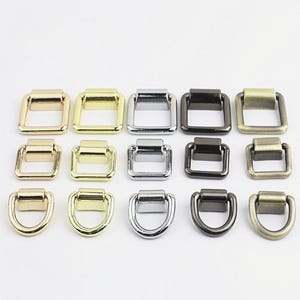Pode incluir: Uma coleção de quinze conectores de metal dispostos em três fileiras. As duas fileiras superiores apresentam alças quadradas, enquanto a fileira inferior consiste em anéis em forma de D. Os acabamentos incluem ouro polido, prata, preto e latão.