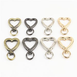 May include: A collection of eight heart-shaped swivel lobster clasps arranged in two rows. The metal clasps feature four distinct finishes: antique brass, gunmetal, silver, and gold, each equipped with a circular ring at the base for attachment.