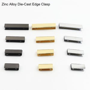 May include: A collection of twelve rectangular zinc alloy die-cast edge clasps arranged in three columns by color: matte black, polished gold, and polished silver. Each clasp features a U-shaped profile with two small circular holes on the side.