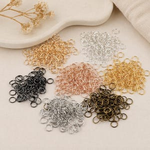 May include: A collection of small metal jump rings arranged in seven distinct piles. The colours include gold, silver, rose gold, black, and antique bronze. These circular jewellery findings are displayed in separate clusters on a neutral surface.