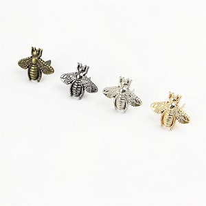 May include: Four metal bee-shaped pins or buttons arranged in a row. Each bee features detailed wings and a segmented body. The set includes one antique brass, one gunmetal, one silver-toned, and one bright gold-toned finish for each individual bee.