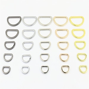 May include: A collection of twenty-five metal D-rings arranged in a grid of five rows and five columns. The rings are organized by color, featuring shades of gunmetal, silver, nickel, rose gold, and gold, with each column decreasing in size.