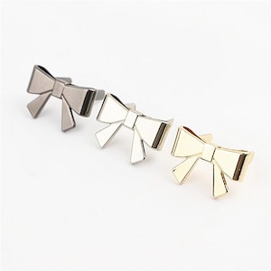 May include: Three metal bow-shaped fasteners arranged in a diagonal row. The first bow is dark gunmetal grey, the second is polished silver, and the third is bright gold. Each bow features a central knot and two downward-pointing ribbon tails.