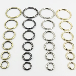 Könnte beinhalten: Eine Sammlung von kreisförmigen Metall-Federringen, angeordnet in vier vertikalen Spalten nach Farbe, darunter Antikmessing, Gunmetal, Silber und Goldtöne. Jeder Ring verfügt über einen Klappmechanismus zum sicheren Befestigen.