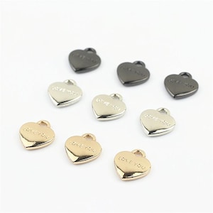 Puede incluir: Una colección de nueve dijes de metal en forma de corazón en tonos negro, plateado y dorado. Cada dije cuenta con una pequeña anilla en la parte superior para su sujeción y el texto LOVE YOU grabado claramente en la parte frontal de la pieza.
