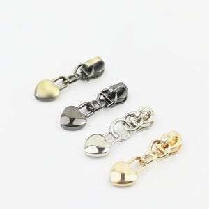 May include: Four metal zip pulls arranged in a diagonal line, each featuring a heart-shaped charm attached by a small circular ring. The set includes finishes in antique brass, gunmetal, polished silver, and bright gold tones for sewing projects.
