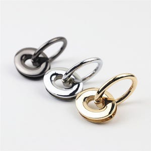 Könnte beinhalten: Drei Metall-Taschenverbinder in Gunmetal-, Silber- und Goldtönen. Jedes Teil besteht aus einer flachen, kreisförmigen Basis mit glatter, polierter Oberfläche und einem beweglichen, runden Ring in der Mitte, der zur Befestigung von Riemen dient.