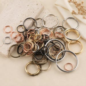 May include: A collection of metal split rings in various sizes and finishes, including silver, gold, copper, and dark bronze tones. These circular hardware components are designed for keychains and organisation, featuring a coiled wire construction.