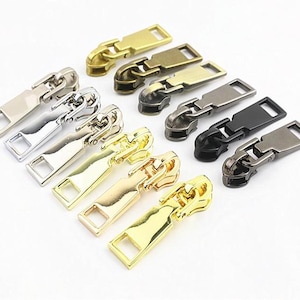 Könnte beinhalten: Eine Sammlung von zehn Reißverschluss-Anhängern aus Metall, angeordnet in zwei Reihen. Das Set enthält verschiedene metallische Oberflächen wie poliertes Gold, Silber, Mattschwarz, Gunmetal und Antikbronze, jeweils mit einer rechteckigen Zuglasche.