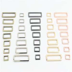 May include: A collection of rectangular and square metal loops in various finishes including antique brass, silver, gunmetal, gold, and rose gold. These hardware components are arranged in columns, showcasing different sizes and metallic tones.