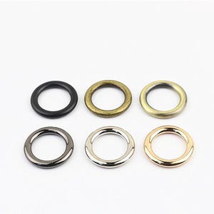 May include: Six circular metal rings arranged in two rows of three. The top row features a matte black ring, an antique brass ring, and a brushed gold ring. The bottom row includes a gunmetal ring, a polished silver ring, and a rose gold ring.
