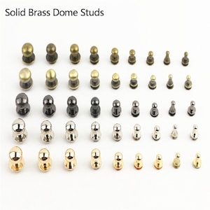 Pode incluir: Uma coleção de tachas de cúpula de latão maciço dispostas em cinco linhas horizontais por cor e tamanho. O texto Solid Brass Dome Studs aparece no topo. As tachas apresentam topos arredondados e acabamentos metálicos variados, incluindo latão antigo, bronze escuro, prata polida e tons dourados brilhantes.