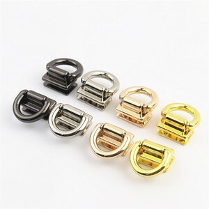 Puede incluir: Un conjunto de ocho conectores metálicos en forma de D dispuestos en dos filas. Las piezas de herraje cuentan con un bucle semicircular unido a una base rectangular, disponibles en acabados metálicos de color bronce, plata y oro.