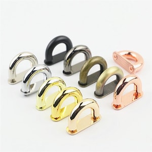 May include: A collection of ten metal D-rings in various finishes, including polished silver, matte black, gunmetal, antique brass, brushed gold, and rose gold. Each D-ring features a curved top loop attached to a flat, rectangular base plate.